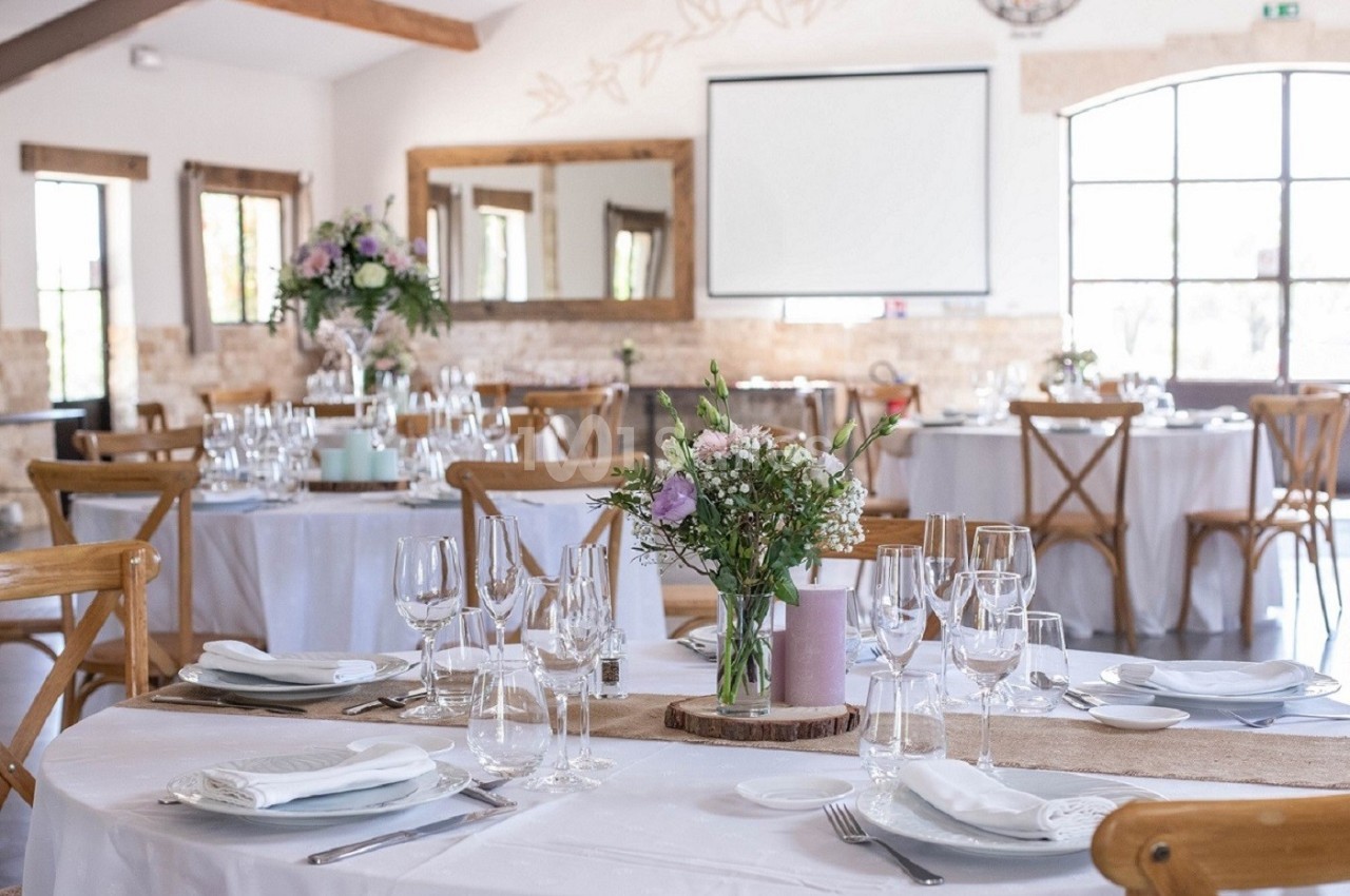 Salle de réception lumineuse avec tables décorées de fleurs, nappes blanches et vaisselle élégante.