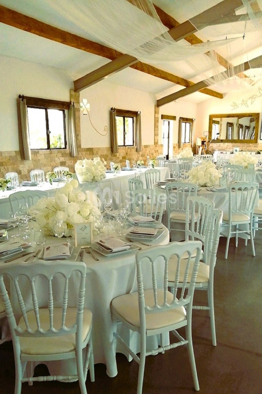 Salle de réception décorée avec des tables rondes, nappes blanches, chaises assorties et centres de table fleuris.