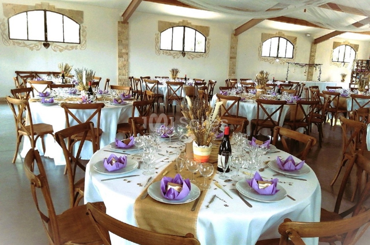 Salle de réception décorée avec des tables rondes, nappes blanches, serviettes violettes et centre de table floral.