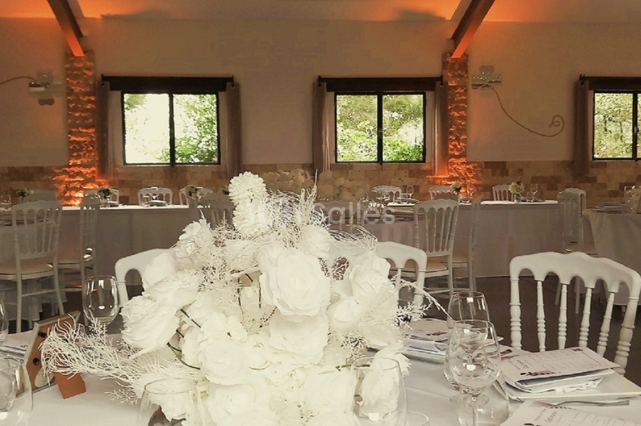Salle de réception décorée avec des tables rondes, nappes blanches, bouquets de fleurs et éclairage chaleureux.