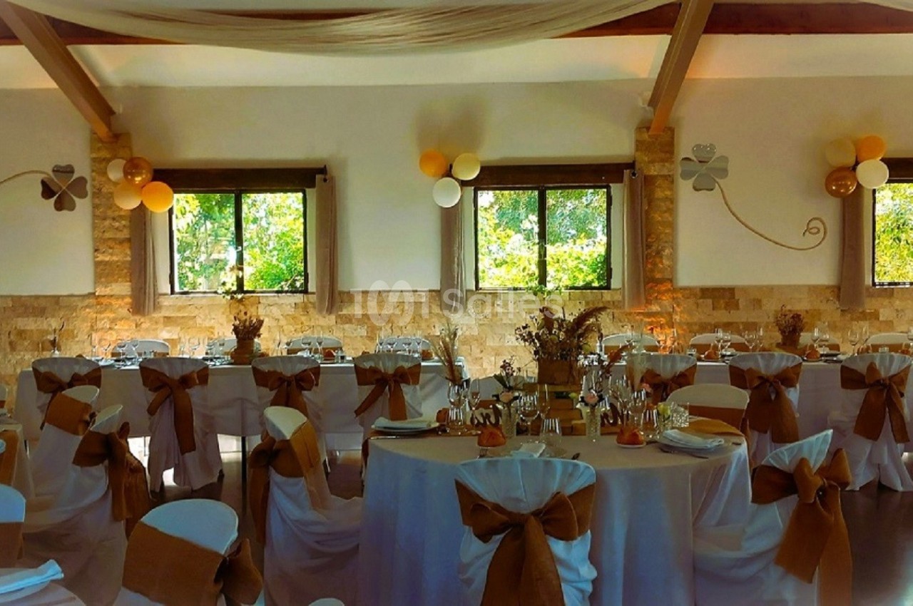 Salle de réception décorée avec des tables rondes, nappes blanches et nœuds marron sur les chaises, éclairée par des…