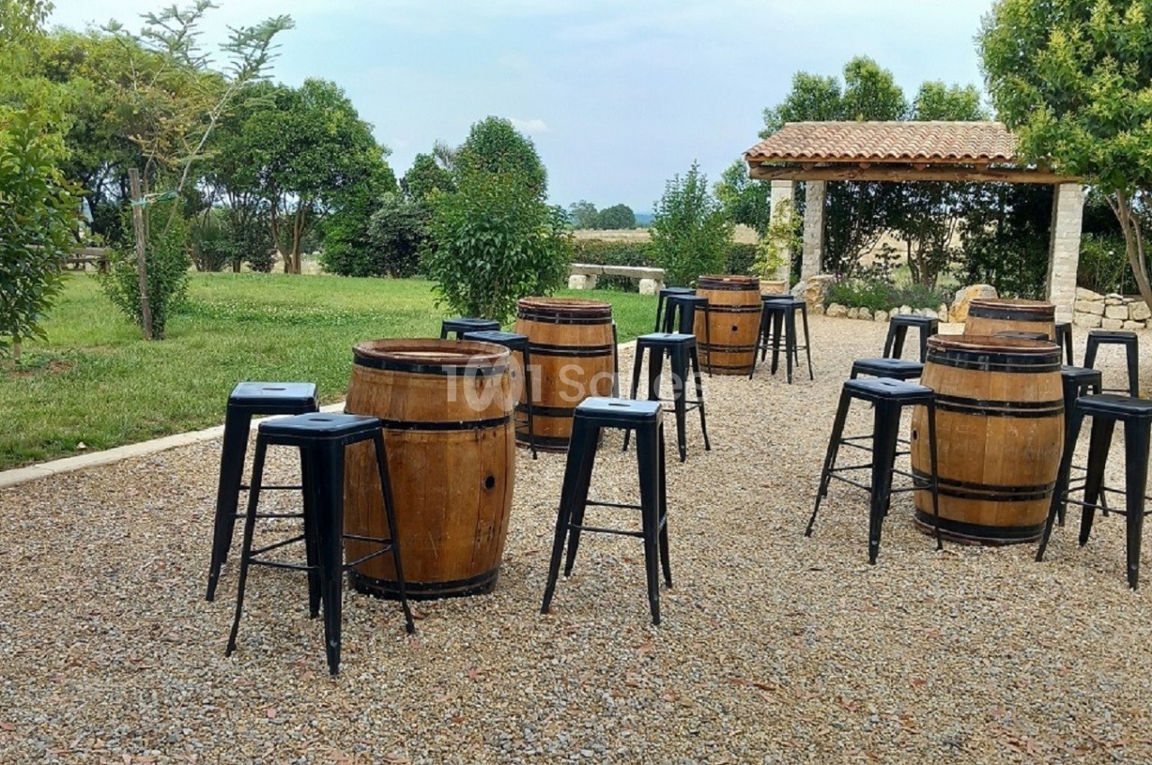 Barriques en bois utilisées comme tables, entourées de tabourets noirs, disposées sur une terrasse en gravier avec un jardin…