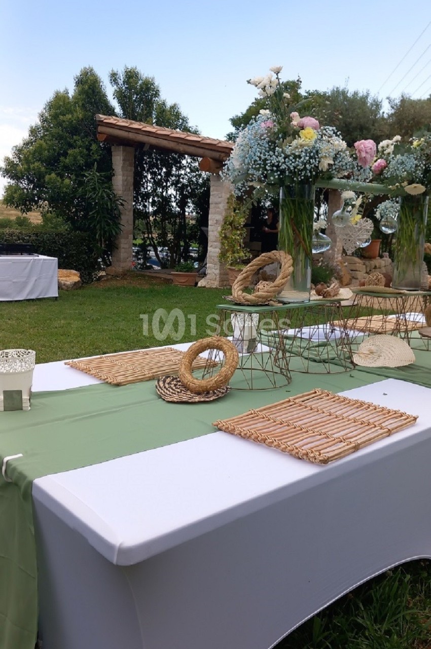 Table décorée en extérieur avec nappes blanches, chemins de table verts, fleurs et éléments en osier dans un jardin.