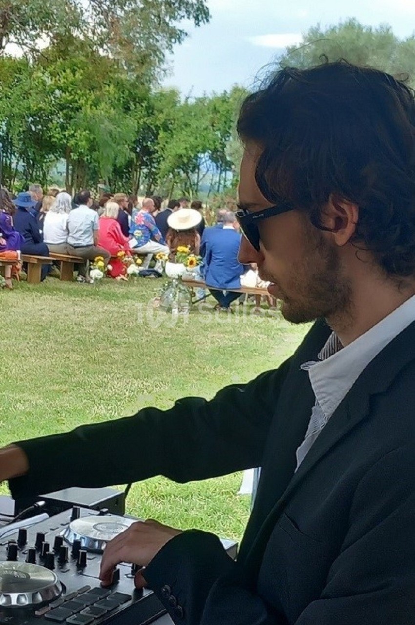 Un DJ mixant en plein air lors d'un événement avec des invités assis sur des bancs dans un cadre verdoyant.
