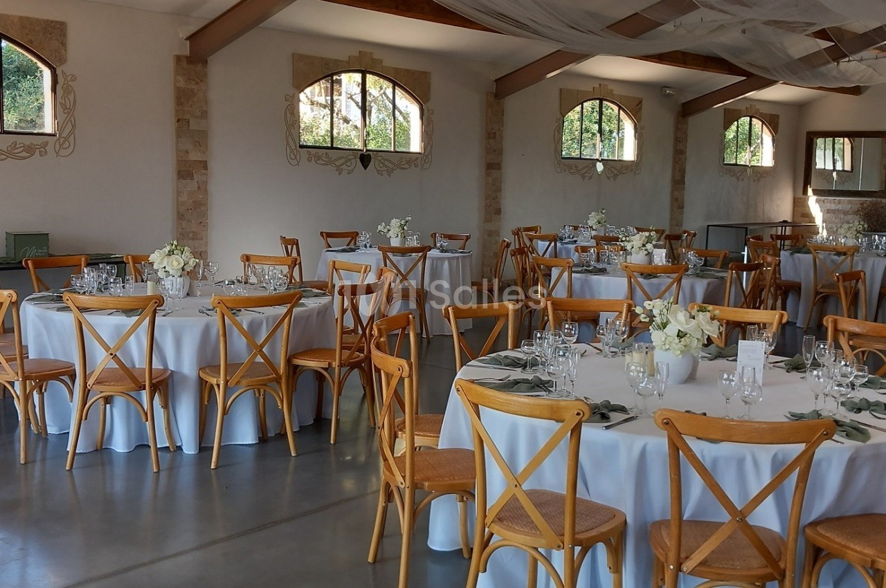 Salle de réception lumineuse avec tables rondes dressées, chaises en bois et décorations florales blanches.