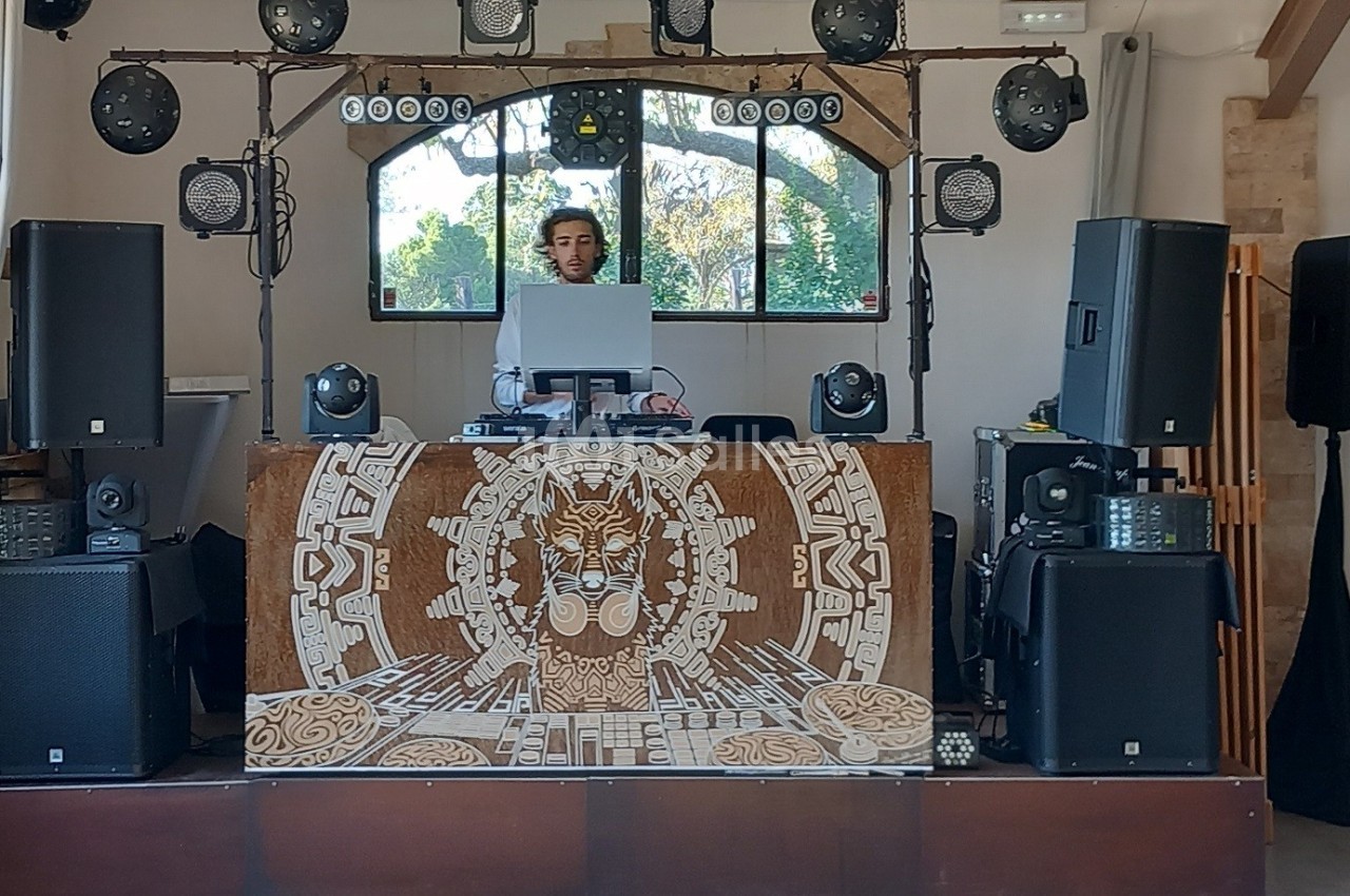 Un DJ mixe derrière une table ornée de motifs artistiques, entouré d'enceintes et d'équipements lumineux.