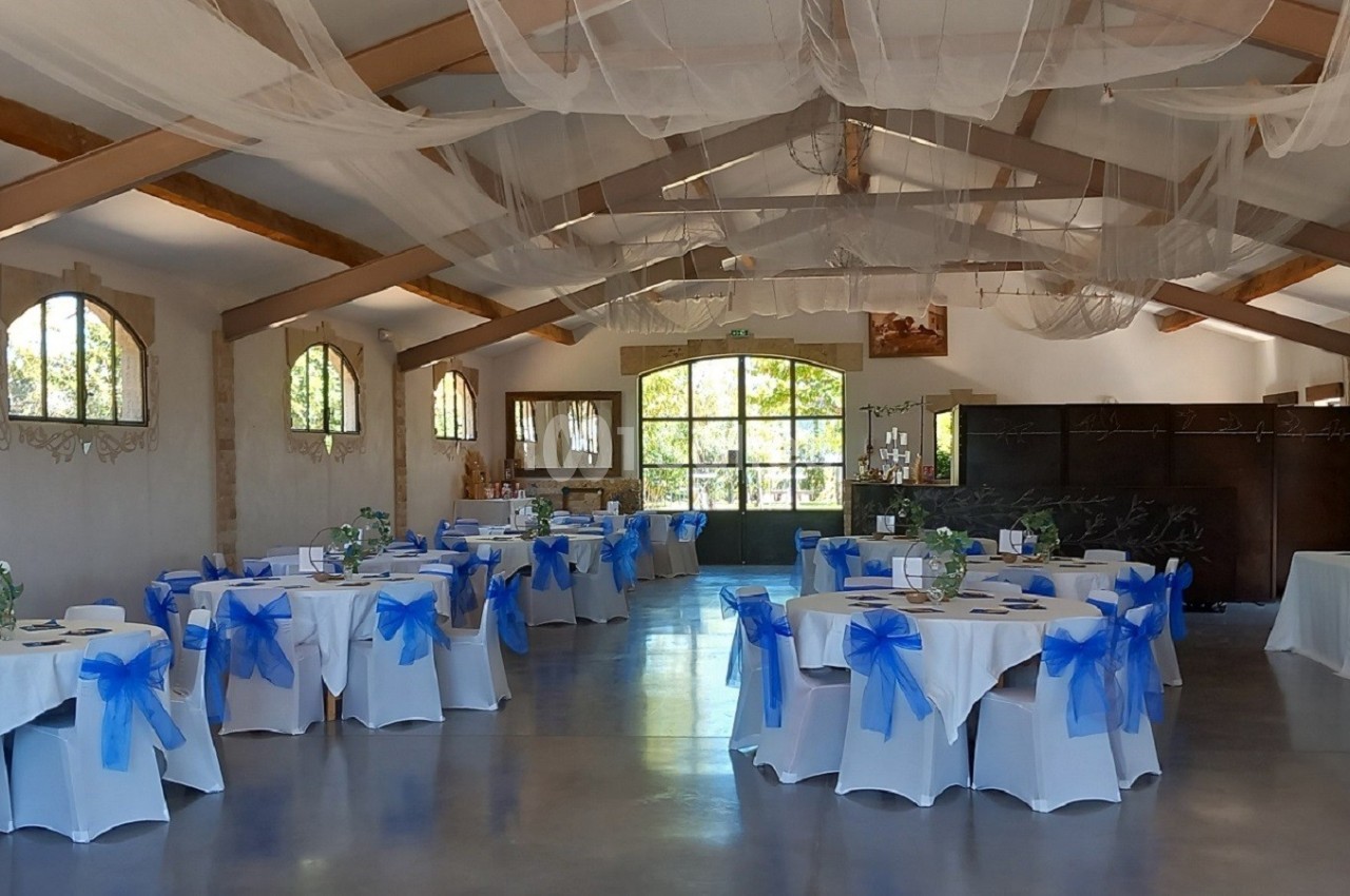 Salle de réception décorée pour un événement, avec des tables rondes ornées de nappes blanches et nœuds bleus.