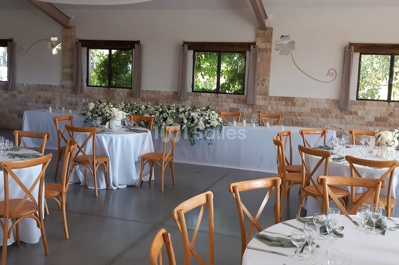 Salle de réception lumineuse avec tables rondes dressées, chaises en bois et décoration florale blanche et verte.