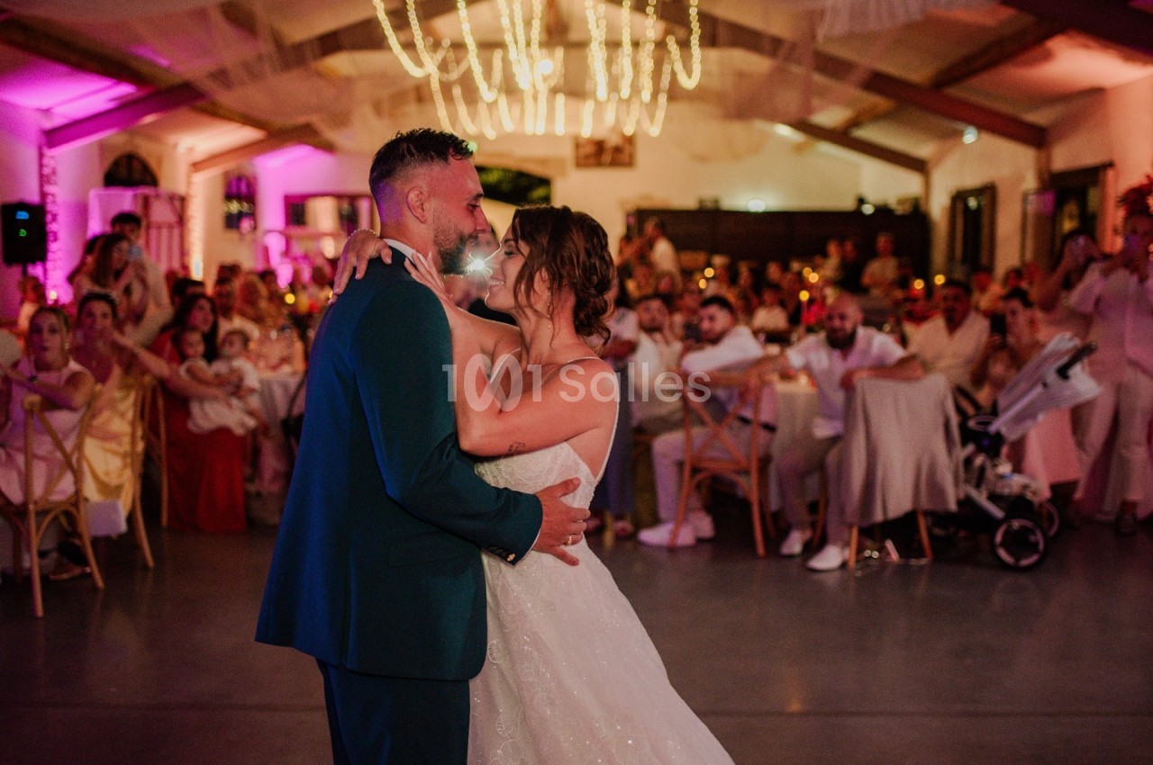 Un couple en tenue de mariage danse sous un éclairage chaleureux dans une salle décorée, entouré d'invités.