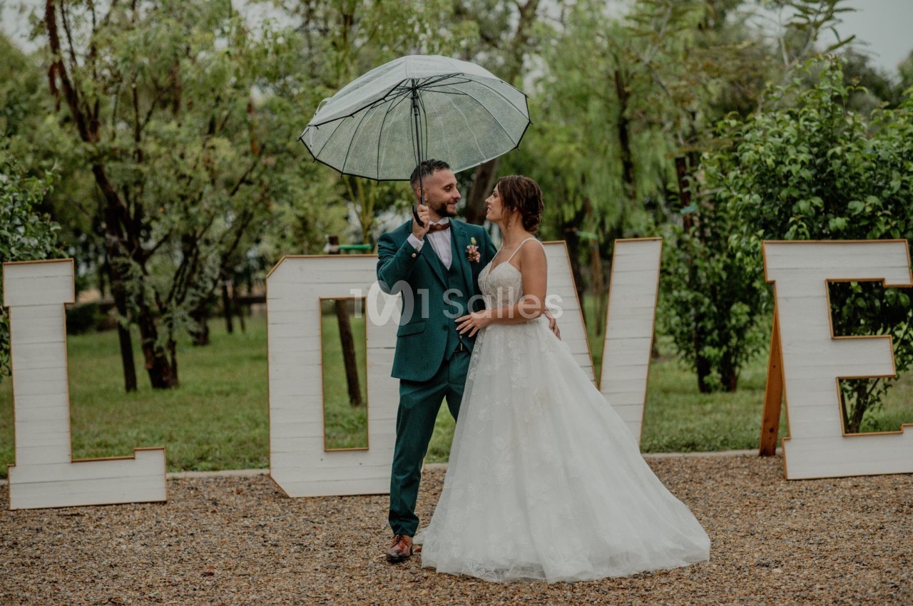 Un couple en tenue de mariage sous un parapluie transparent, devant des lettres formant le mot ’LOVE’ dans un jardin.