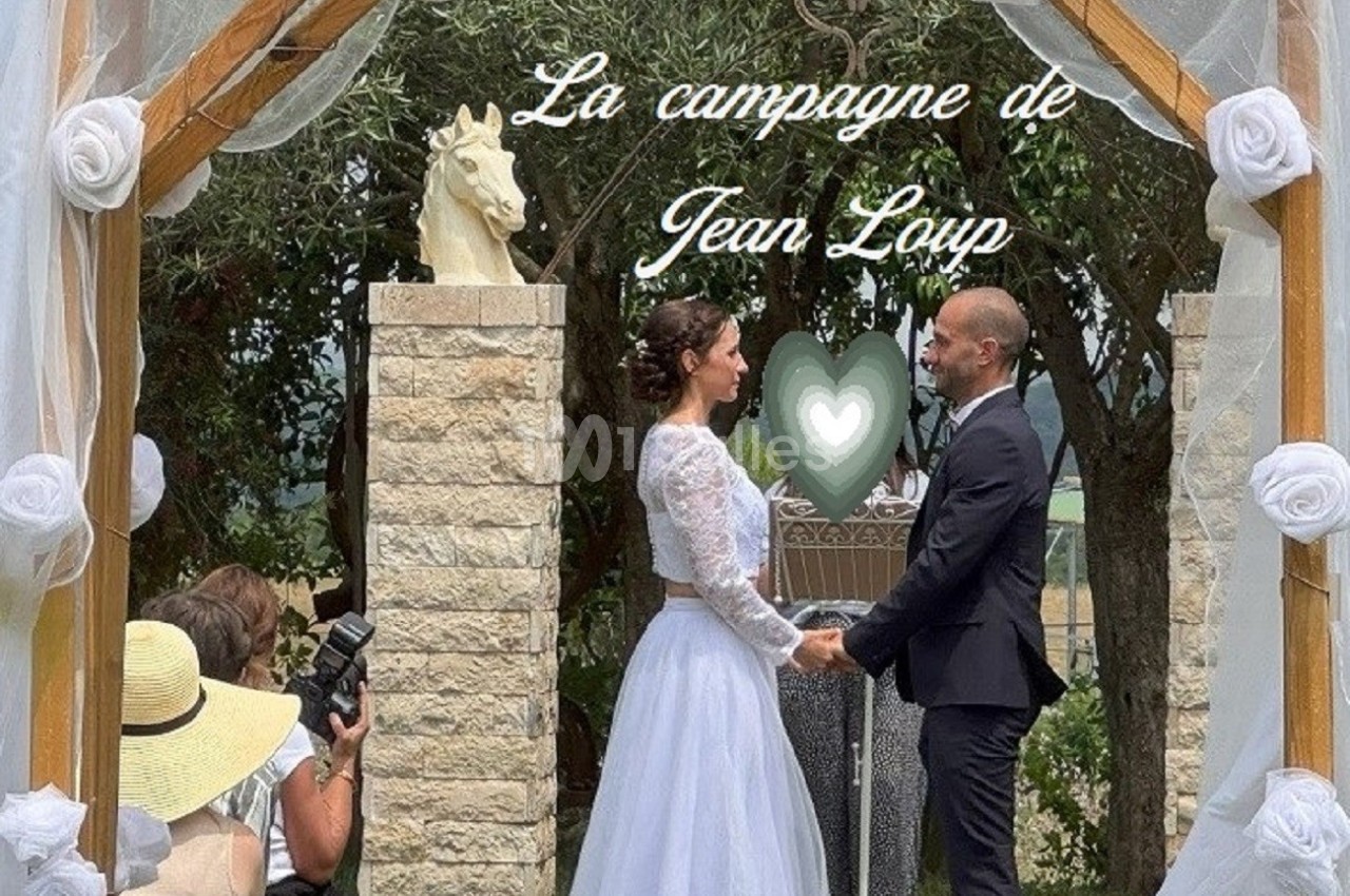 Un couple échange ses vœux sous une arche décorée de voilages et de fleurs, en extérieur, devant des invités.