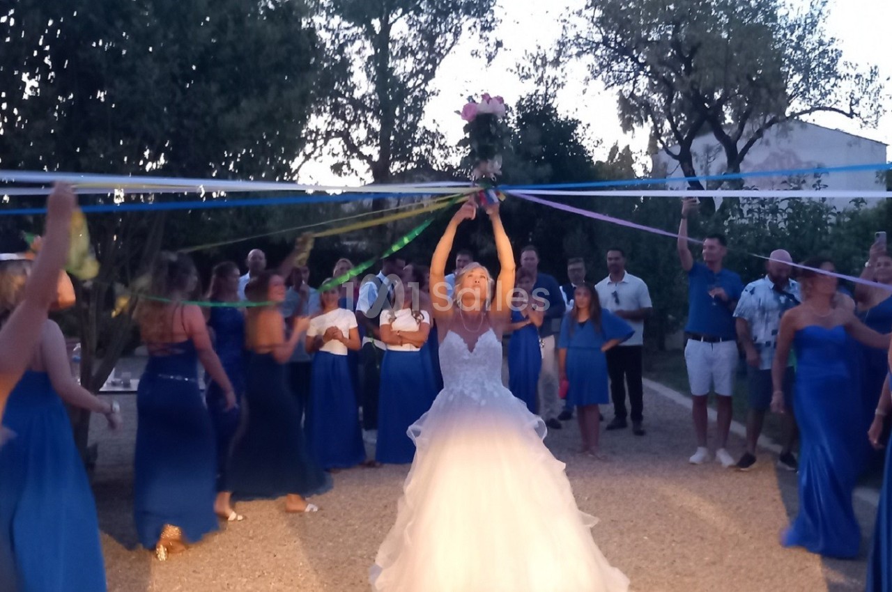 Une mariée lance son bouquet dans les airs, entourée d'invités en tenue bleue dans un jardin en soirée.