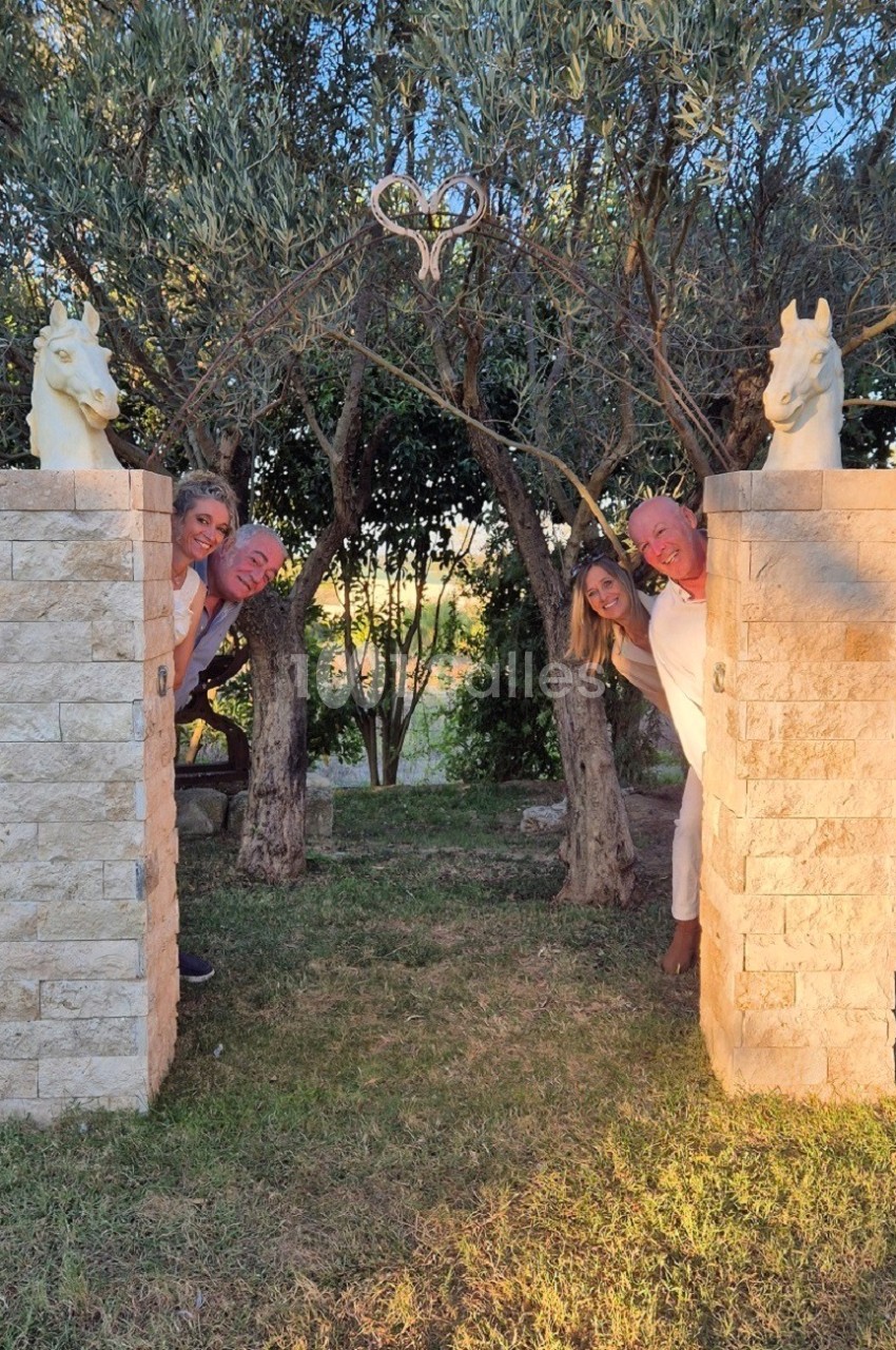 Quatre personnes souriantes se cachent derrière deux piliers en pierre surmontés de statues de chevaux, dans un jardin au…