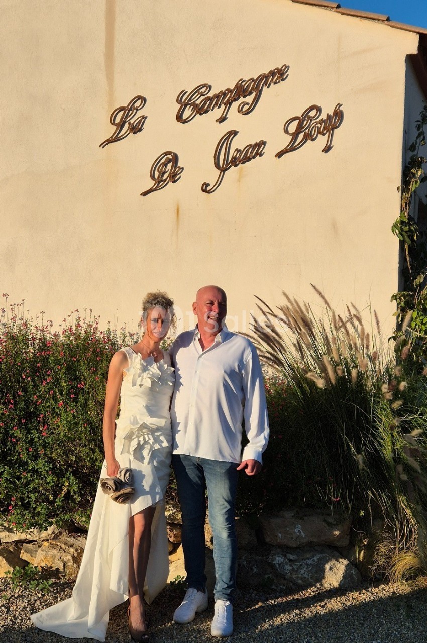 Un couple pose devant un mur beige avec l'inscription ’La Campagne de Jean Loup’, entouré de végétation.