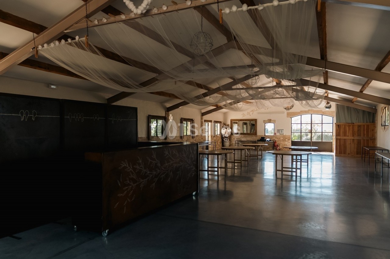 Salle spacieuse avec poutres apparentes, tables et décorations suspendues, éclairée par de grandes fenêtres.