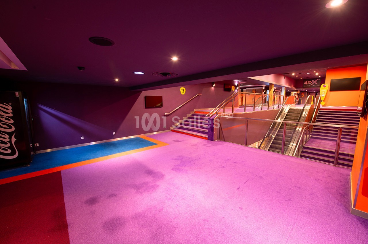 Escalier et escalator dans un hall de cinéma aux couleurs vives avec moquette violette et affichages lumineux.