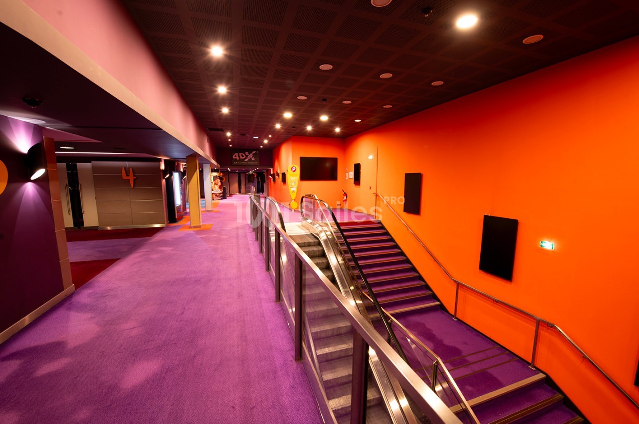 Escalier et couloir d'un cinéma aux murs orange et sol violet, éclairés par des spots au plafond.