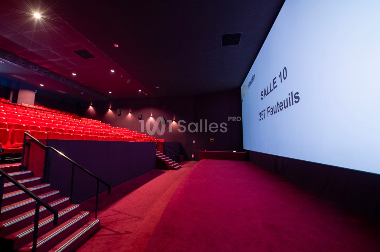 Salle de cinéma vide avec sièges rouges, écran géant affichant ’Salle 10, 257 fauteuils’ et allée centrale.