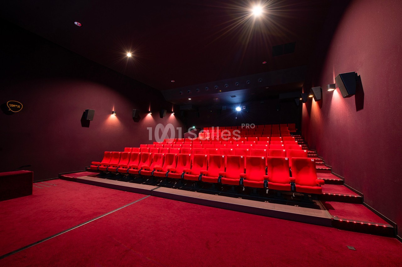 Salle de cinéma vide avec des rangées de sièges rouges, éclairée par des lumières tamisées.