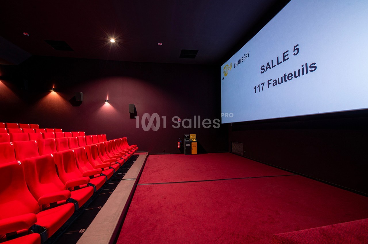 Salle de cinéma vide avec des sièges rouges, un écran affichant ’Salle 5 - 117 Fauteuils’ et un sol recouvert de moquette…