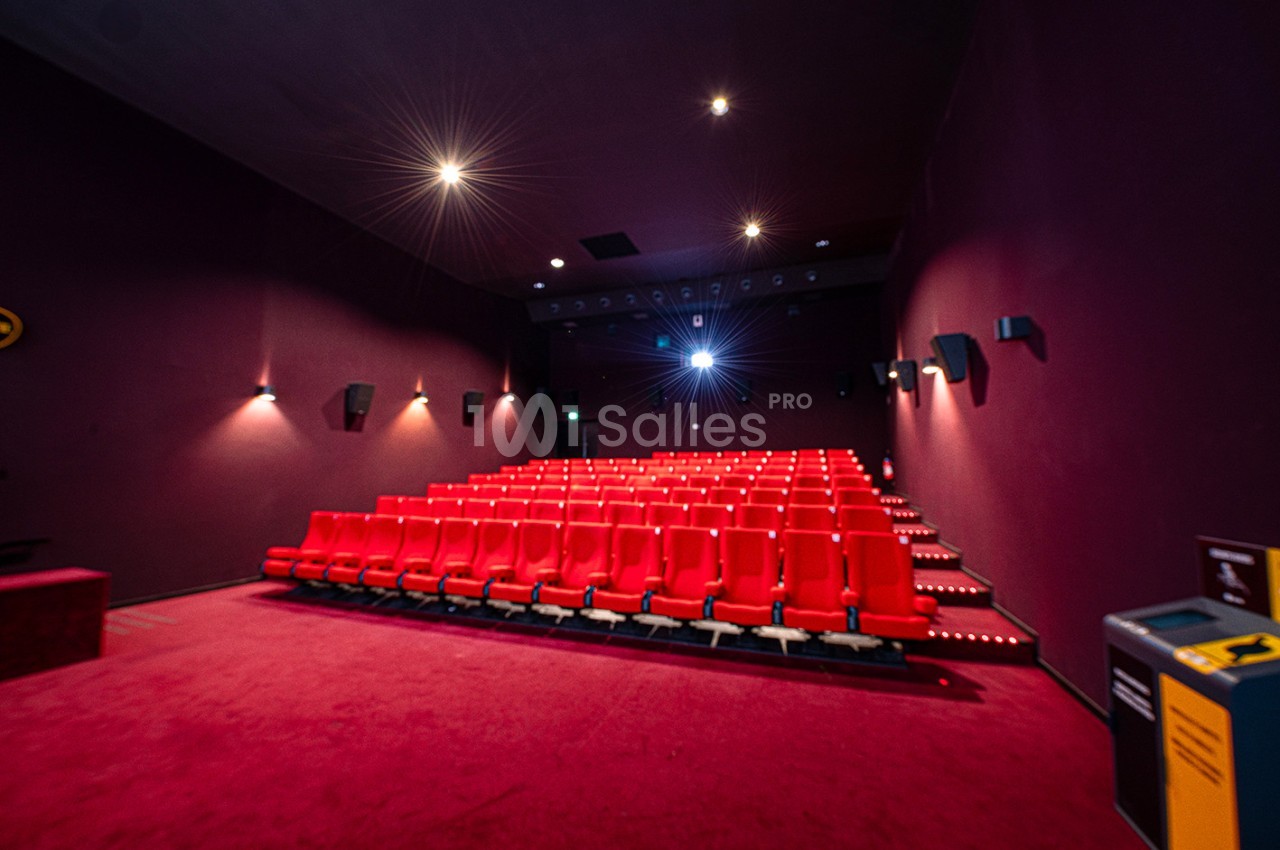 Salle de cinéma vide avec rangées de sièges rouges, éclairage tamisé et projecteur allumé.