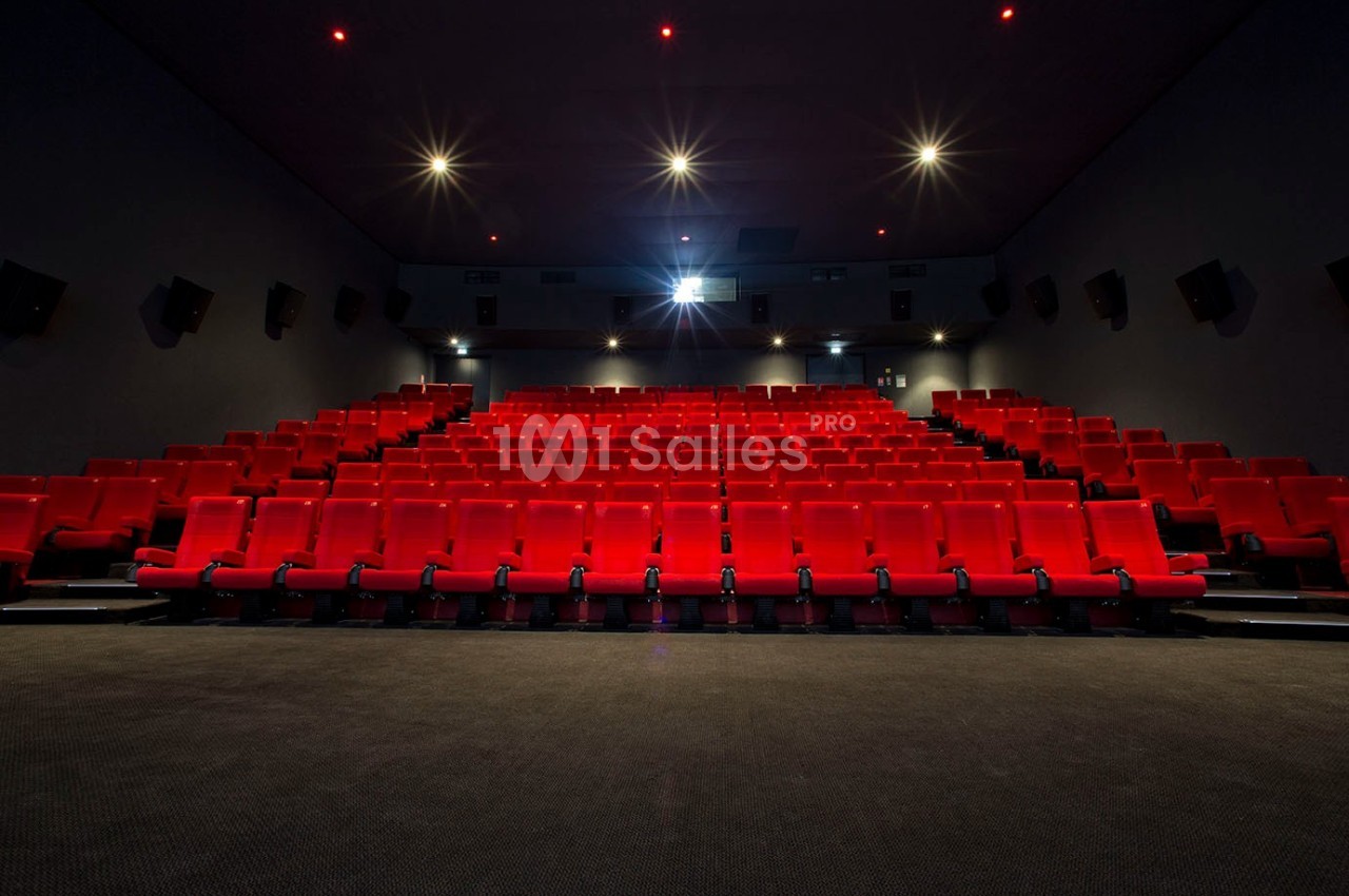 Salle de cinéma vide avec des rangées de sièges rouges et un écran blanc éclairé à l'arrière-plan.