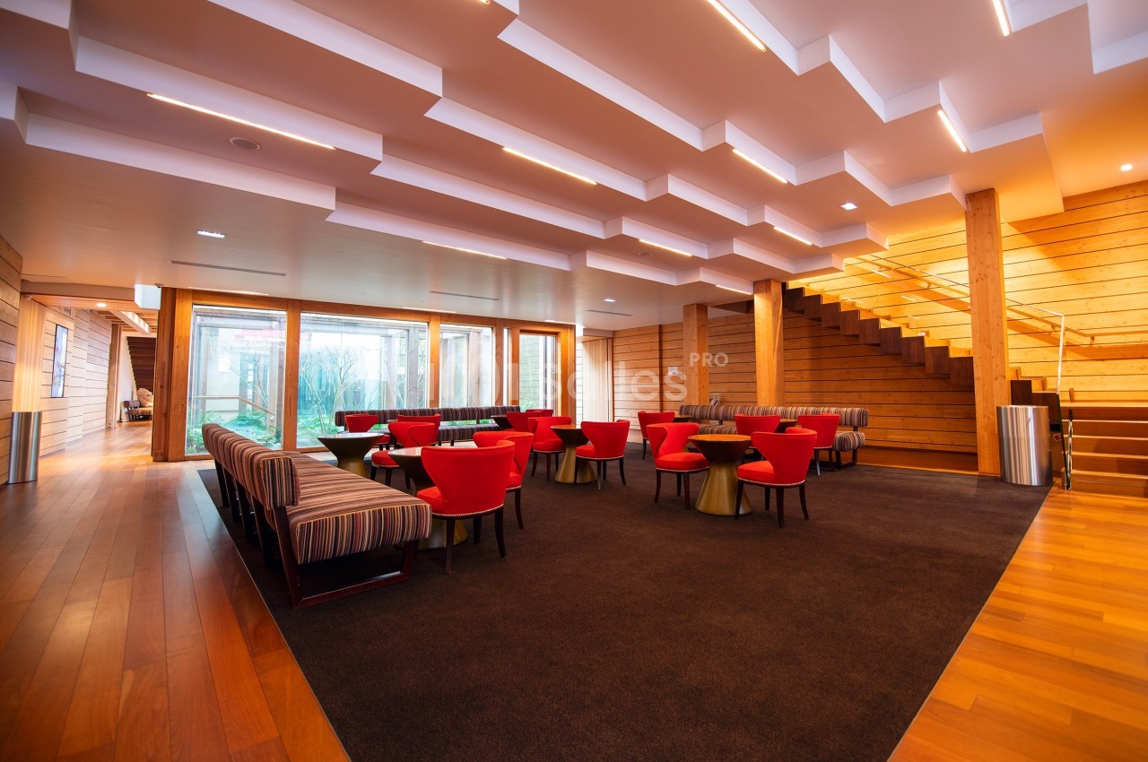 Salle lumineuse avec des fauteuils rouges, des banquettes rayées, un escalier en bois et de grandes baies vitrées.