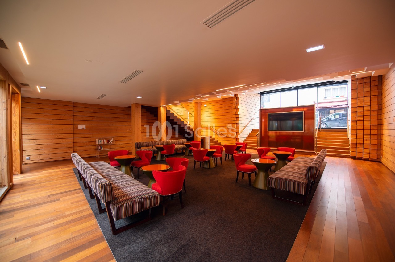 Salle avec mobilier moderne, comprenant des fauteuils rouges, des tables basses et des murs en bois clair.