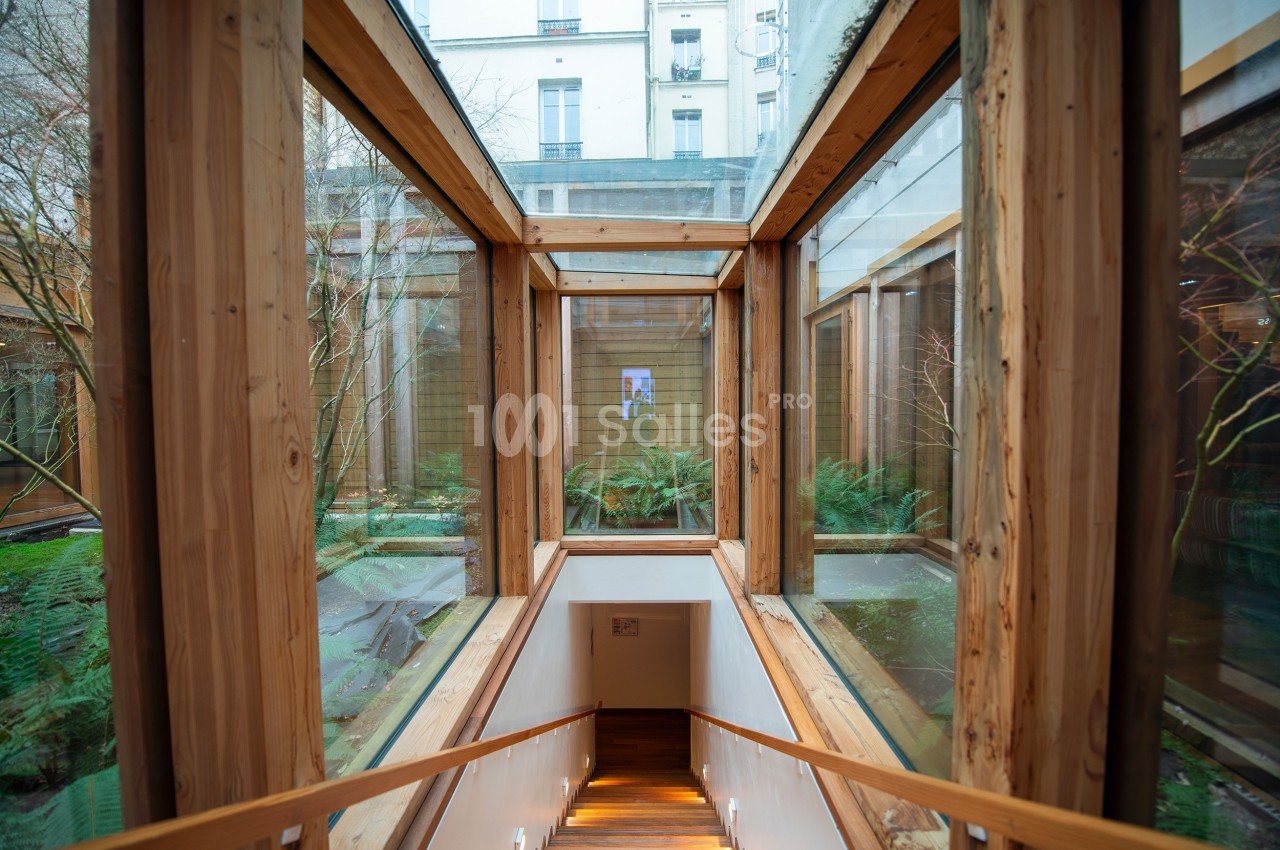 Passage en bois vitré menant à un escalier, avec vue sur un jardin intérieur et des bâtiments en arrière-plan.