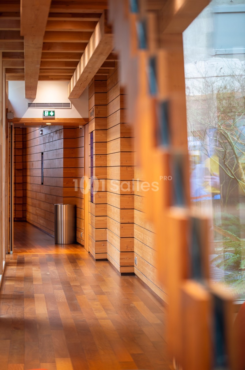 Couloir en bois avec parquet, murs lambrissés, poubelle métallique et lumière naturelle entrant par une grande fenêtre.