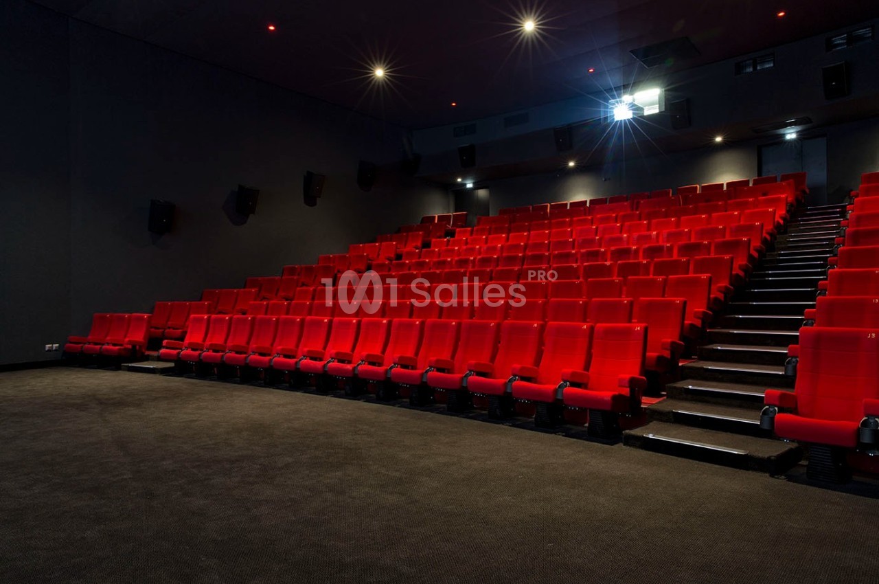 Salle de cinéma vide avec des rangées de sièges rouges et un projecteur allumé.