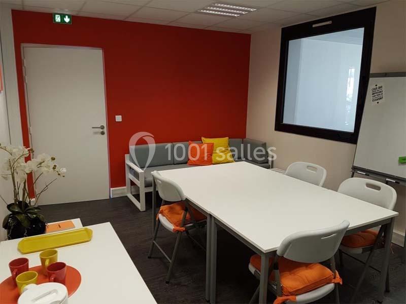 Salle de réunion avec table blanche, chaises pliantes, canapé gris, coussins colorés et mur rouge vif.