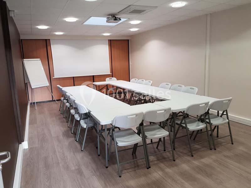 Salle de réunion avec tables blanches disposées en U, chaises pliantes et un paperboard près d'un écran de projection.