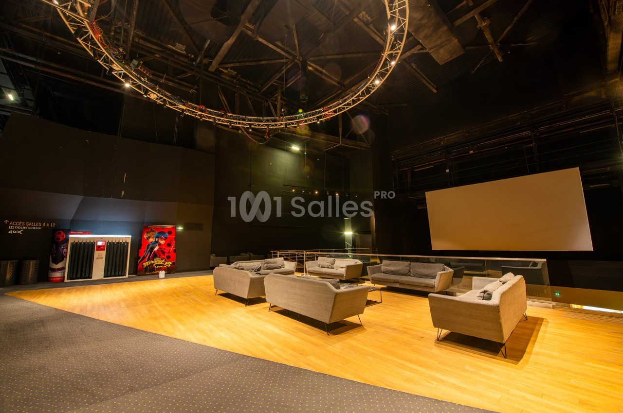 Salle avec des canapés gris, écran blanc, éclairage tamisé et structure métallique suspendue au plafond.