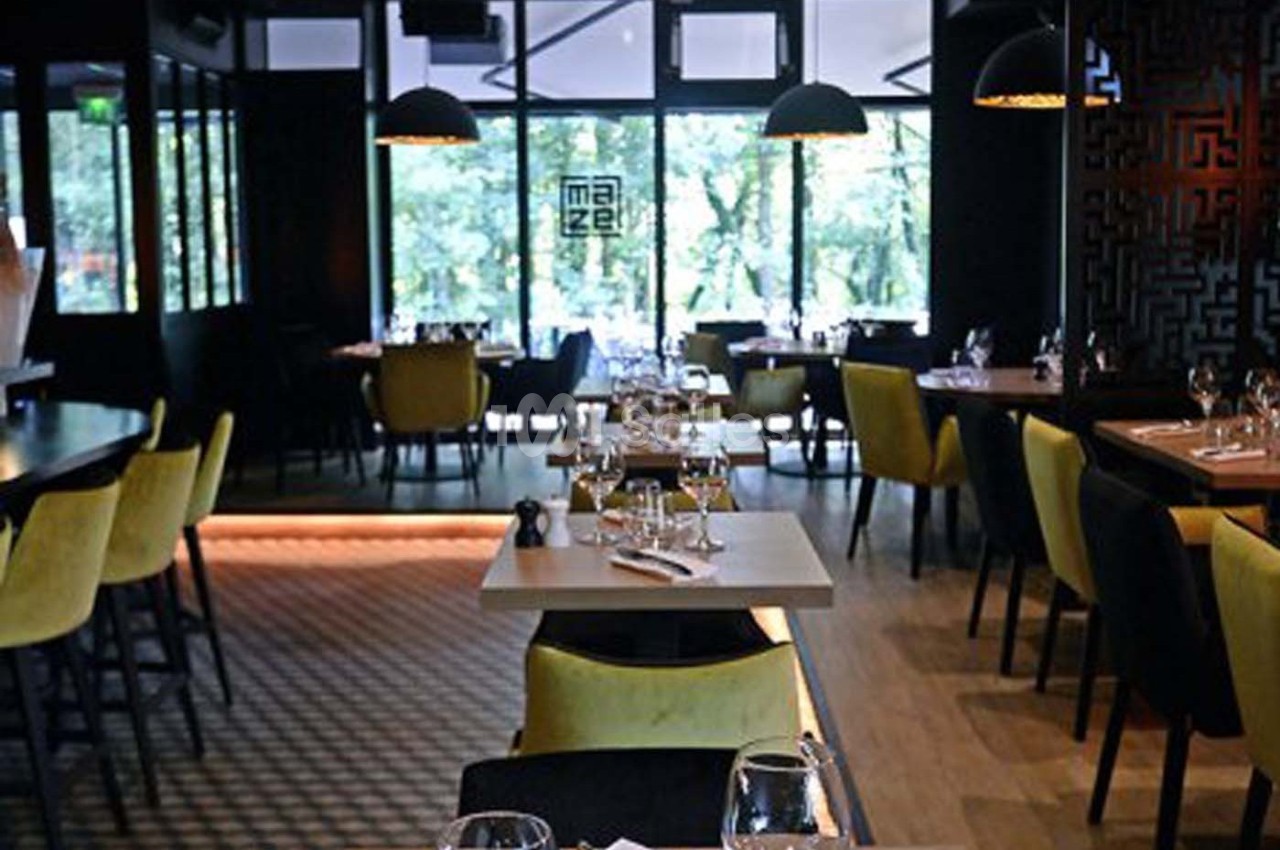 Salle de restaurant moderne avec tables dressées, chaises jaunes et noires, éclairage tamisé et grandes baies vitrées.