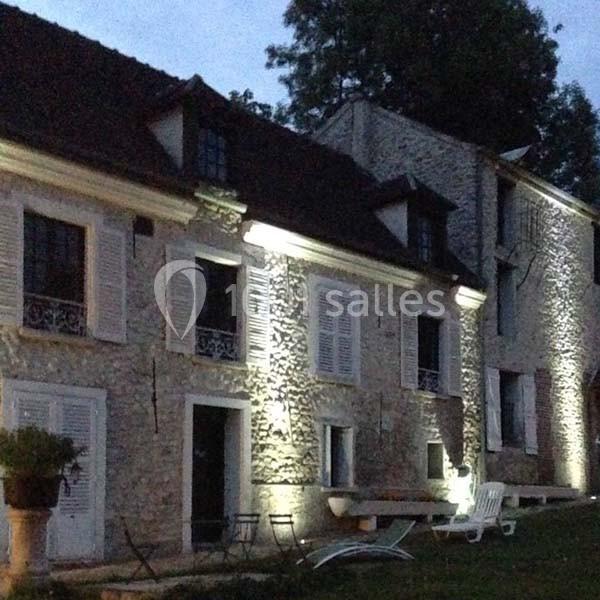 Façade en pierre d'une maison éclairée par des lumières extérieures, avec volets blancs et mobilier de jardin.
