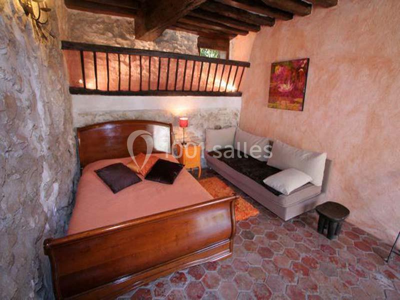 Chambre rustique avec lit en bois, canapé, murs en pierre et poutres apparentes, sol en tomettes rouges.