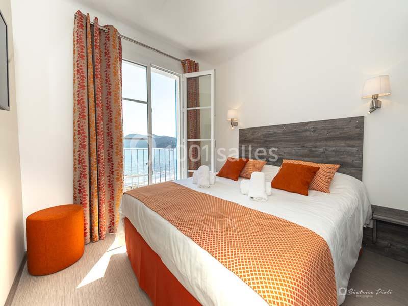 Chambre lumineuse avec lit double, décorée en orange, donnant sur un balcon avec vue sur la mer.
