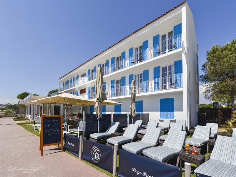 Façade d'un bâtiment blanc à volets bleus avec terrasse aménagée de transats et parasols, entourée de verdure.