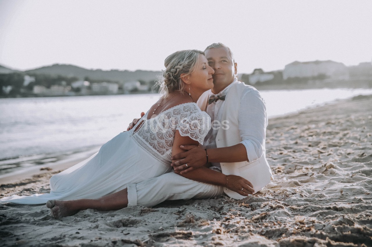 Un couple en tenue de mariage assis sur une plage au coucher du soleil, entouré d'un paysage côtier.