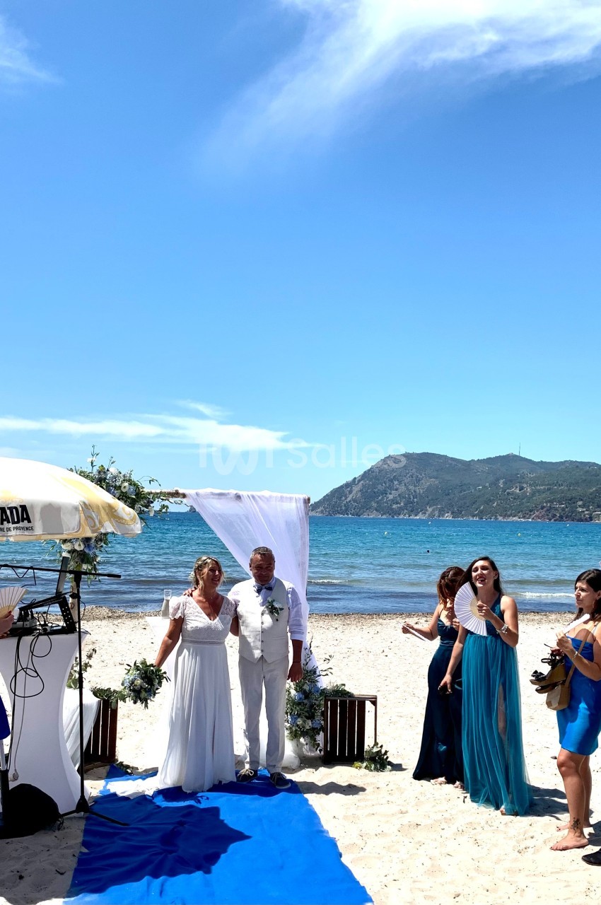Cérémonie de mariage sur une plage, avec un couple sous une arche blanche entouré d'invités et vue sur la mer.