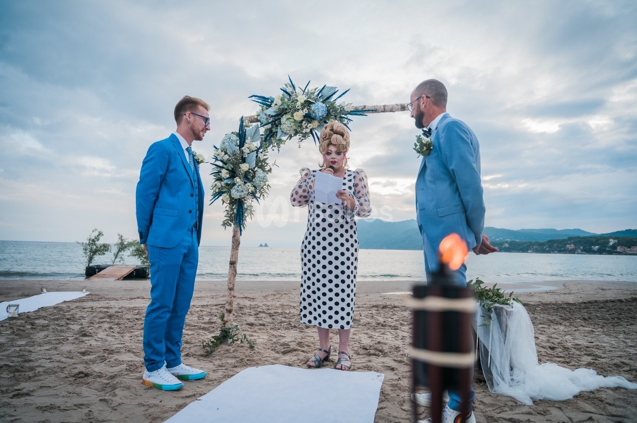 Deux hommes en costume assistent à une cérémonie de mariage sur une plage, officiée par une femme lisant un texte.