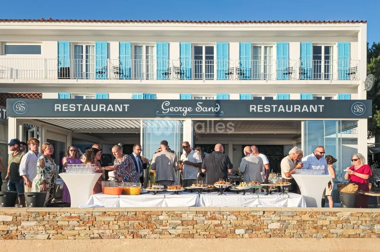 Groupe de personnes rassemblées devant un restaurant avec terrasse, façade blanche et volets bleus.
