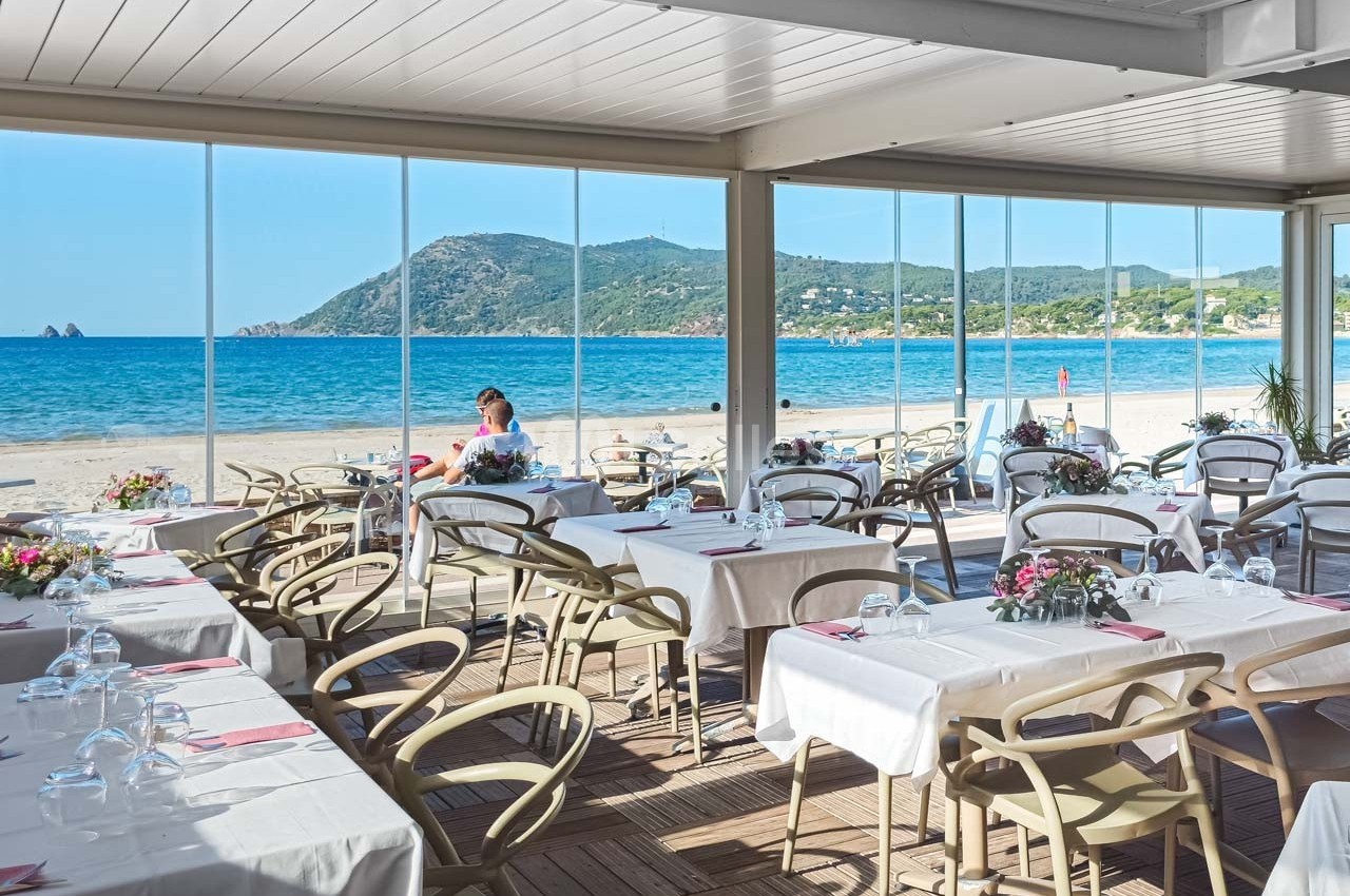 Salle de restaurant lumineuse avec vue panoramique sur une plage, des montagnes et la mer en arrière-plan.