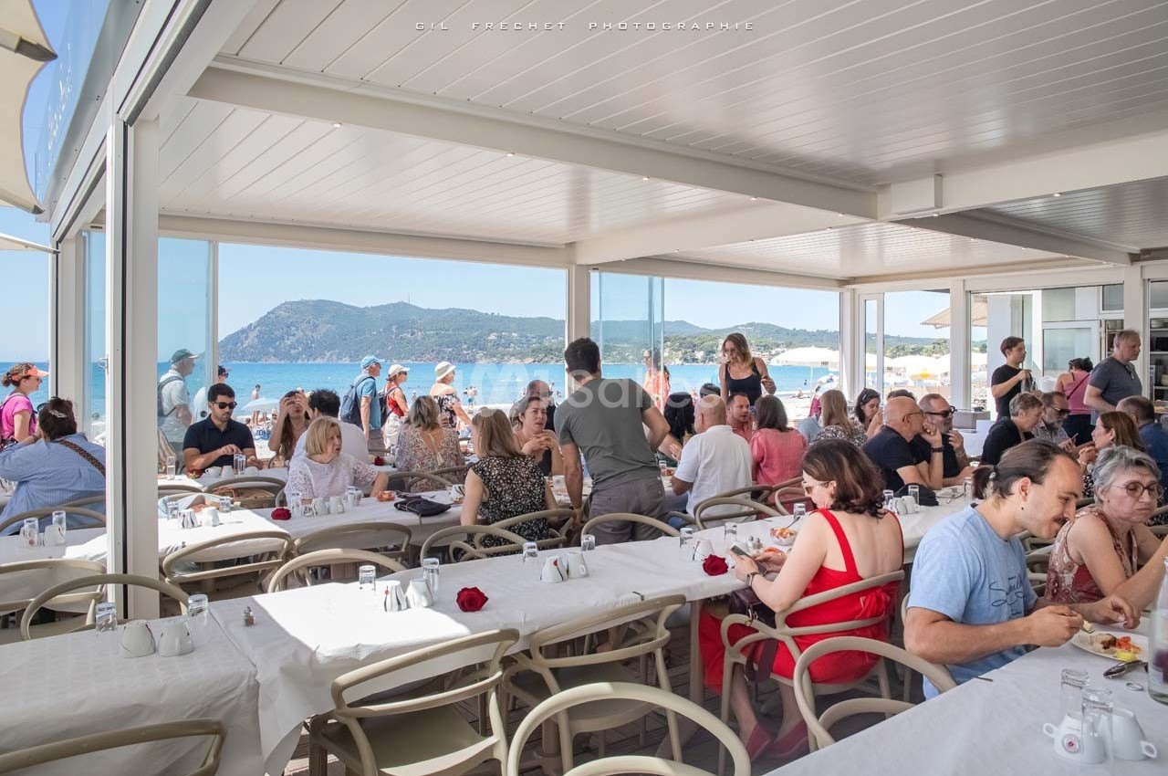 Salle de restaurant en bord de mer avec des clients attablés, vue sur la plage et les montagnes en arrière-plan.