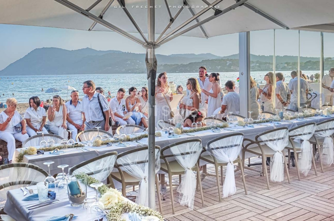 Groupe de personnes en tenue estivale réunies autour de tables décorées, en bord de mer sous un grand parasol.