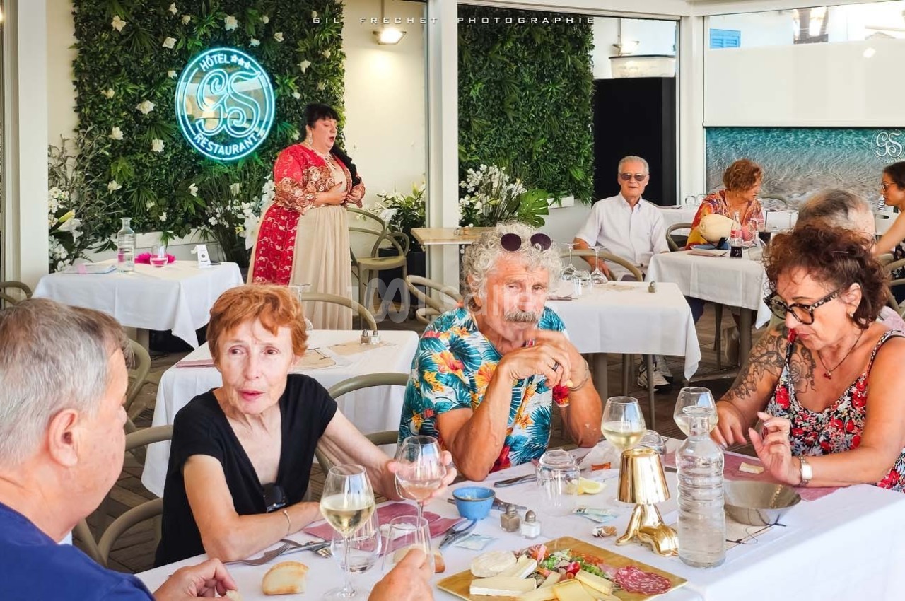 Des personnes d'âges variés discutent autour d'une table dans un restaurant avec terrasse et décoration végétale.