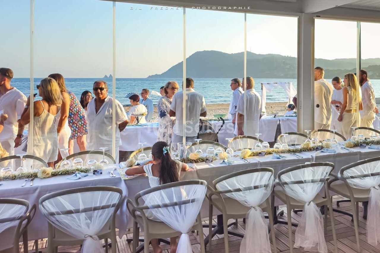 Groupe de personnes en tenue blanche réunies autour de tables décorées, avec vue sur la mer et les montagnes.