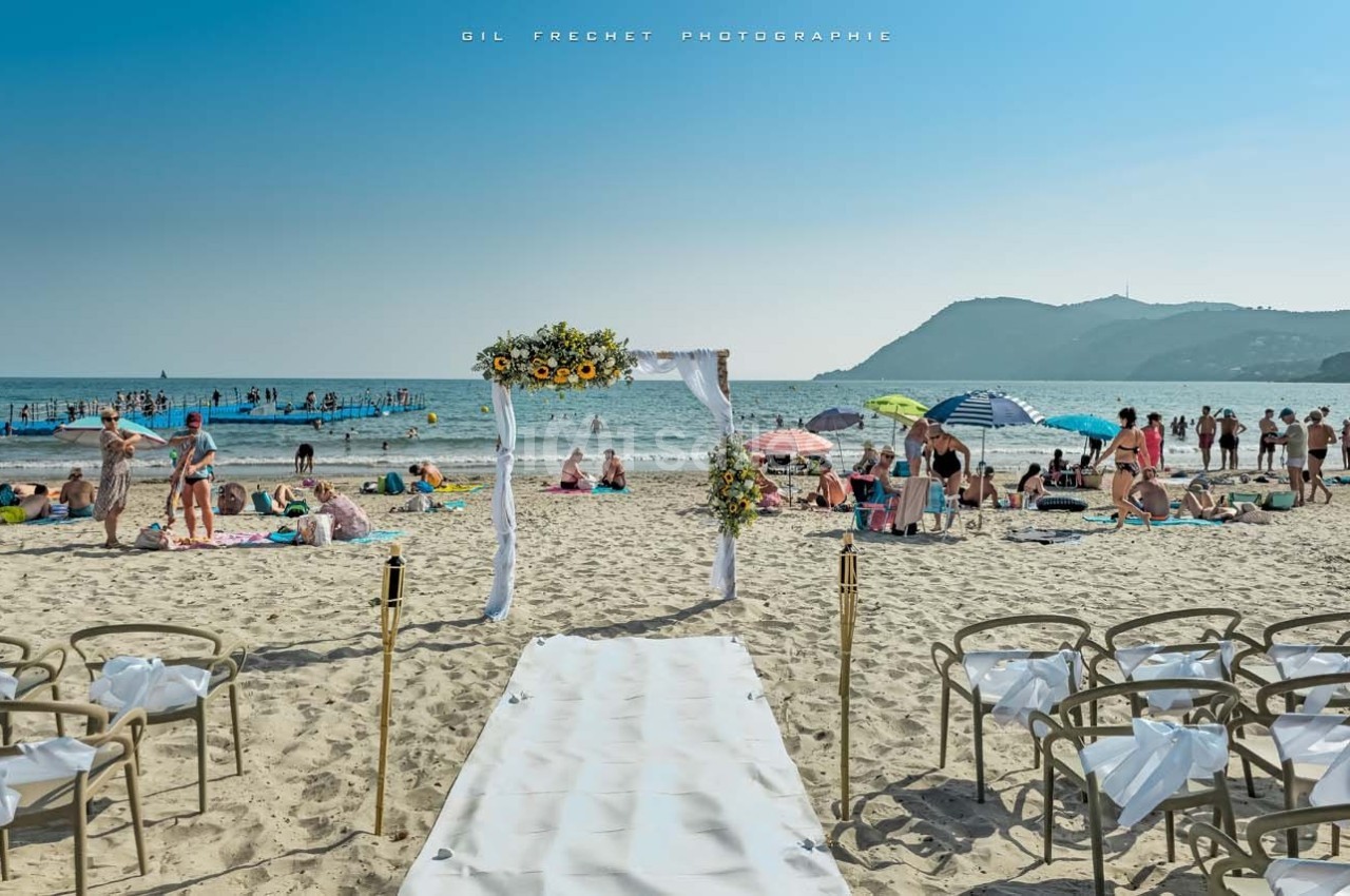 Allée décorée menant à une arche fleurie pour une cérémonie sur une plage animée en bord de mer.