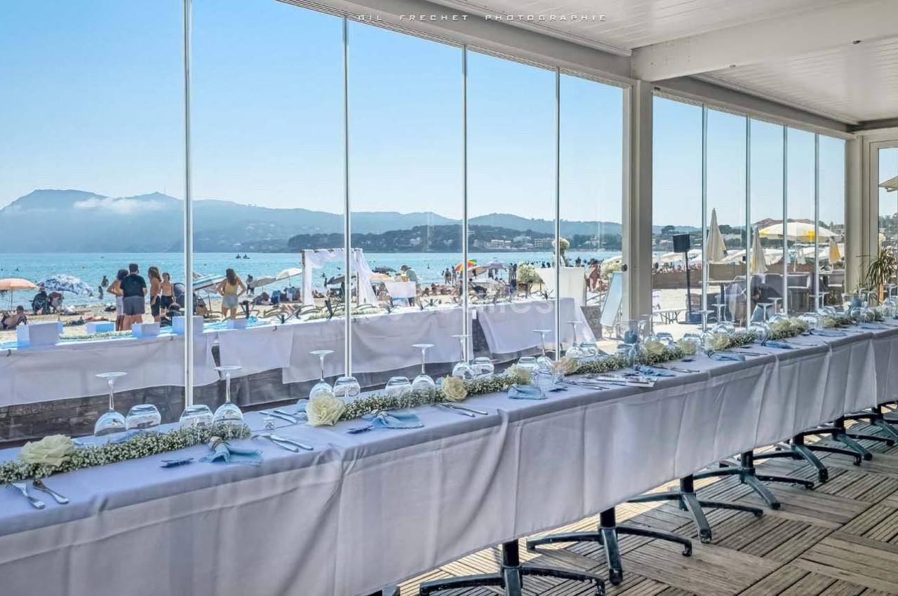 Salle de réception lumineuse avec une longue table dressée, vue sur une plage animée et des montagnes en arrière-plan.